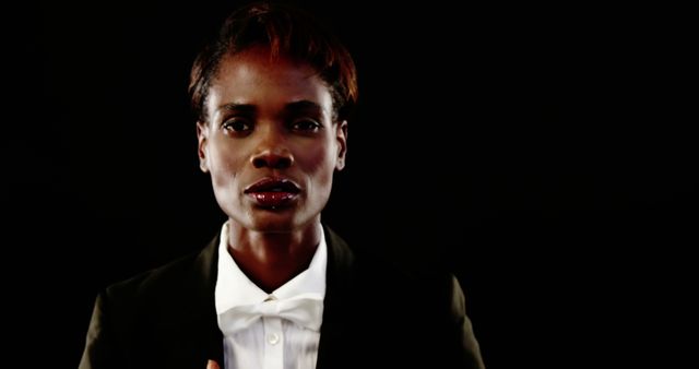 Confident African woman in formal wear with a white bow tie stands against a black background. This image can be used to represent professionalism, leadership, and confidence in corporate or business contexts. Ideal for use in marketing materials, business websites, and leadership articles.