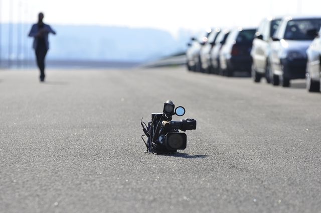 This image features an abandoned video camera lying on asphalt, with a moving runner blurred in the background and parked cars on the side. It can be used in themes about street photography, filming equipment, technology, and urban environments.
