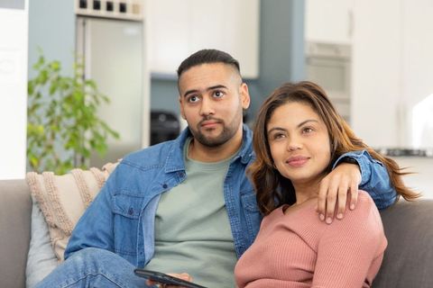 Young couple watching tv and cuddling on sofa in open-plan living room, remote in hand