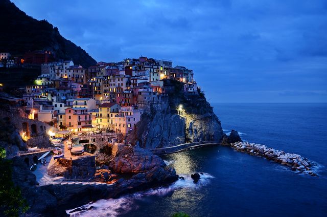 Capturing a picturesque coastal village at dusk, this scene includes charming illuminated buildings nestled on a cliffside. The evening sky casts a tranquil twilight over the ocean, as colorful houses create a striking contrast with the natural surroundings. Ideal for use in travel brochures, tourism websites, and promotional materials for Mediterranean destinations, this image encapsulates the romantic allure and unique architecture of Italy's Cinque Terre. Perfect for adding a serene yet vibrant visual element to any project.
