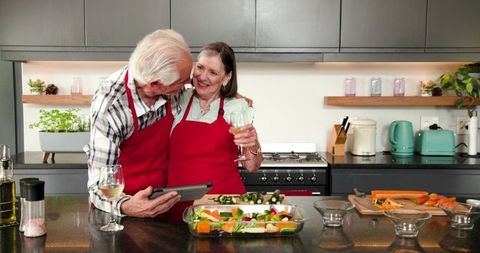 Senior couple cooking together while holding tablet and wine glass