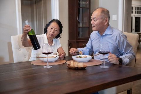 Senior asian couple enjoying cozy home dinner with wine and pasta at wooden table