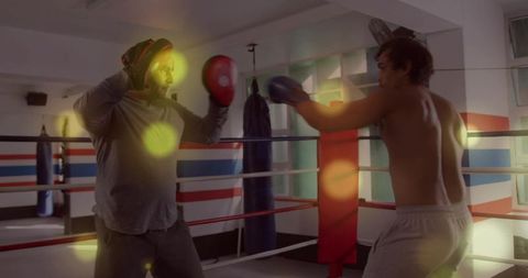 Coach holding focus mitts while shirtless boxer throwing punch in boxing ring with lens flare