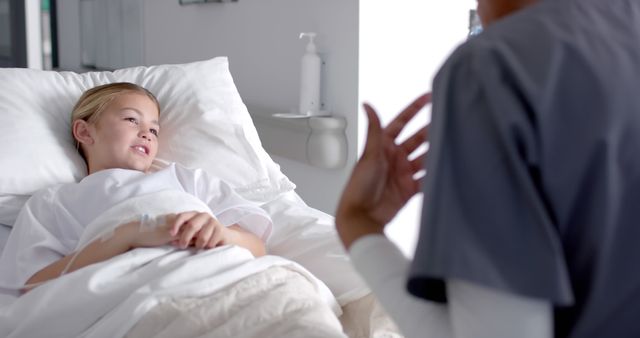 Young girl lying in a hospital bed, having a conversation with a nurse in scrubs. The child appears to be in good spirits, indicating a supportive and positive healthcare environment. Useful for depicting pediatric care, nurse-patient interaction, hospital wellness, and medical treatment.