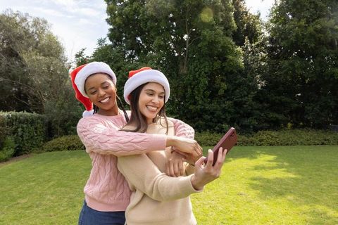 Diverse women wearing santa hats hugging and taking smartphone selfie in sunlit garden