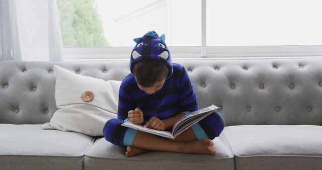 Young child enjoys reading on a couch, wearing a blue dinosaur-themed outfit. Bright natural light from window. Ideal for themes of childhood, imagination, indoor activities or cozy home environments. Perfect for educational content, parenting blogs or advertisements featuring home activities.