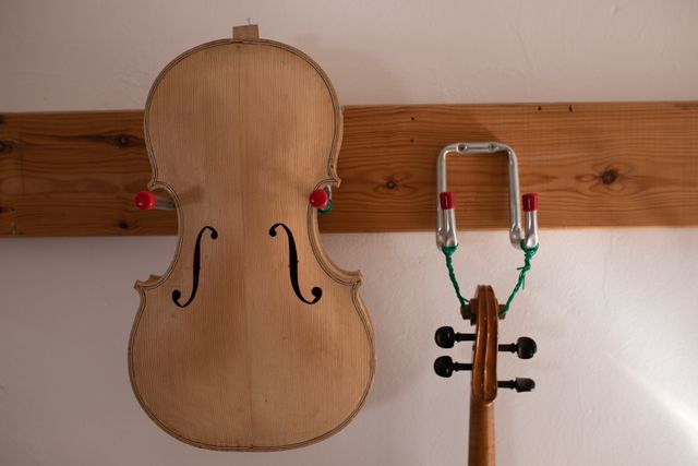 Unfinished violin body and separate neck hanging on wall in luthier's workshop. Sunlight illuminates the workspace, highlighting the craftsmanship involved in creating musical instruments. Ideal for use in articles about traditional craftsmanship, music instrument making, or artisan workshops.
