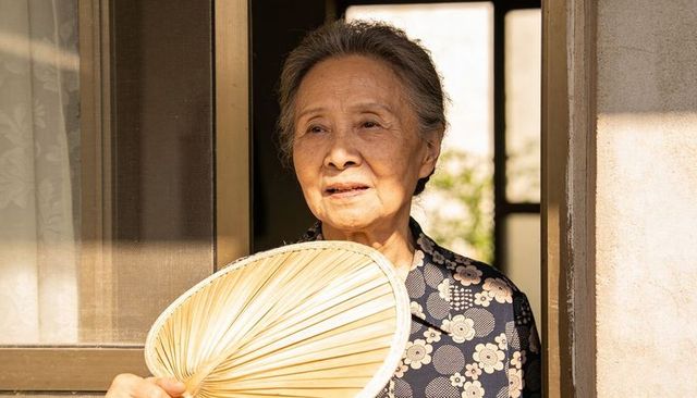 Senior asian woman holding woven bamboo fan at home doorway bathed in golden sunlight