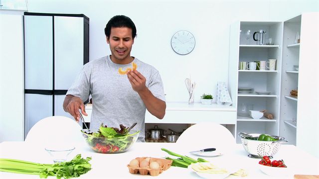 Middle-aged man multitasking and snacking while preparing a fresh salad in a bright, modern kitchen. Useful for advertisements promoting healthy lifestyle recipes, domestic kitchen appliances, and cooking tutorials. Highlights the enjoyment of homemade meal preparation and healthy eating habits.