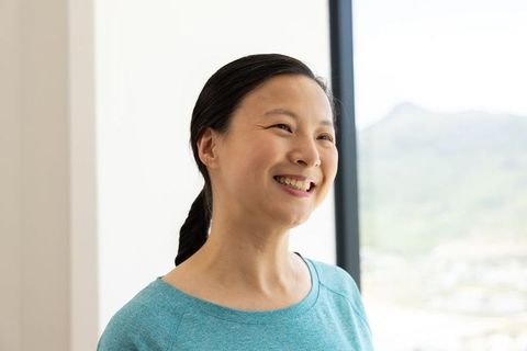 Smiling asian mature woman in teal top by sunlit window minimalist indoor portrait