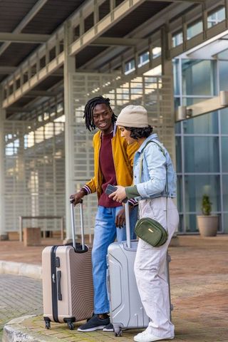 Young diverse couple standing and talking with rolling suitcases at transport terminal