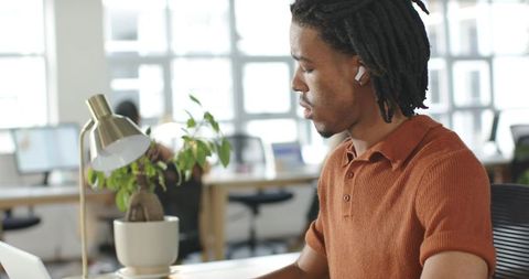 African American man working at desk with laptop and earbuds focusing in bright office