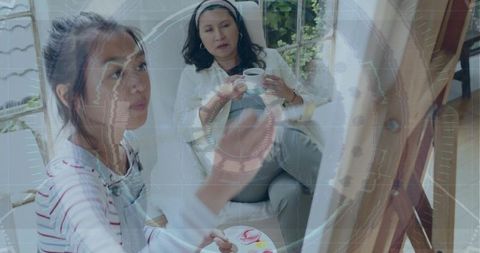 Young artist painting at easel in sunroom while companion sipping coffee and watching