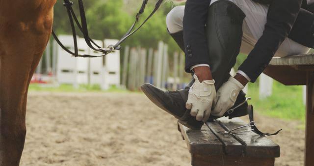This image captures an equestrian preparing for a horseback riding session. The scene highlights the rider's attire, including gloves and boots, emphasizing the sport's preparation process. Ideal for use in equestrian sports promotions, horseback riding tutorials, or content related to outdoor activities and riding gear.