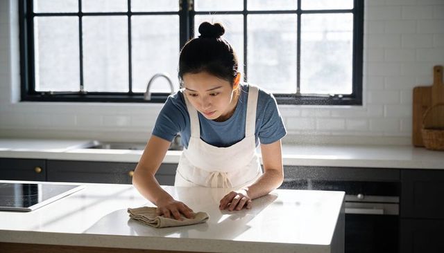 Sunlit asian woman cleaning modern minimalist kitchen island countertop in white apron