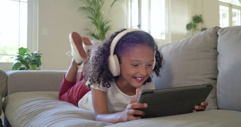 Girl watching tablet while listening with headphones on sofa smiling and relaxed in bright lounge