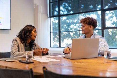 Colleagues discussing charts at conference table with laptop and presentation screen