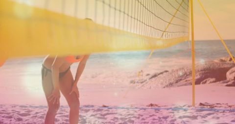 Female beach volleyball player leaning with hands on knees under yellow net at sunset