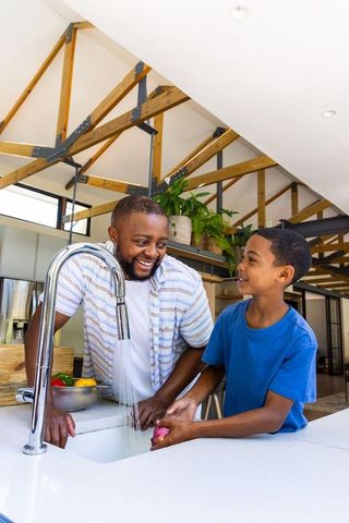 African american father teaching son washing veggies at modern kitchen sink