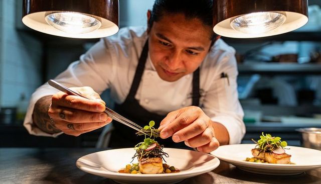 Chef plating microgreens on gourmet scallop dish under pass lamps, precision garnishing