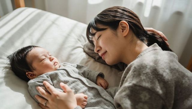 Mother cuddling sleeping newborn on bed in soft daylight, intimate newborn bonding moment