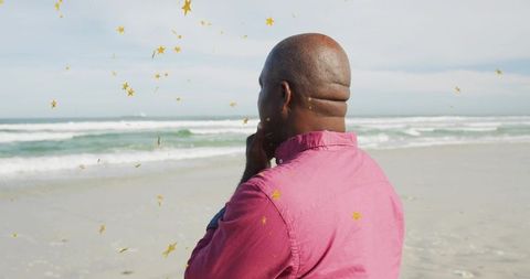 Senior man standing facing ocean on sandy beach wearing pink shirt with gold star overlays