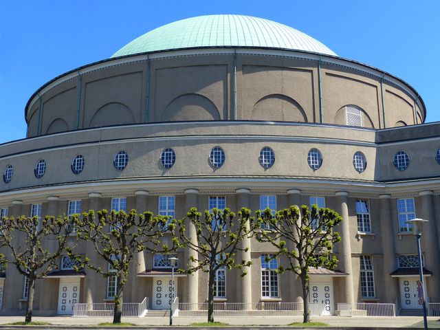 Exterior of a historic building with a large dome and rows of pruned trees against a clear blue sky. The architecture features symmetrical design and classic elements. Suitable for use in articles about architecture, history, and urban landscapes. Ideal as a background for websites or editorial layouts.