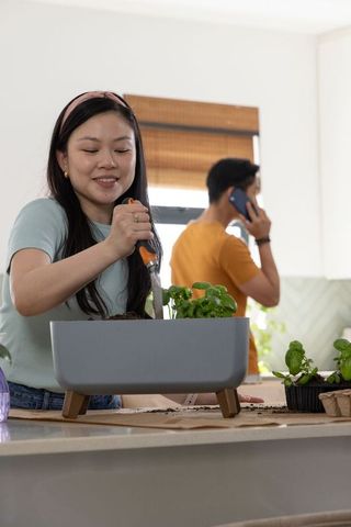 Asian couple planting herbs in kitchen with modern gray planter and hand trowel