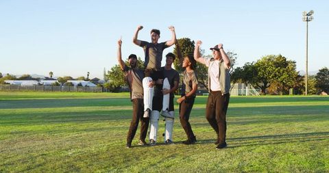 Cricket team celebrating by lifting batsman on grassy field during golden hour victory