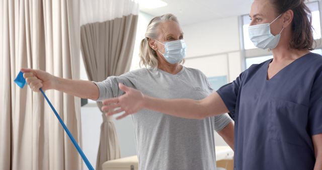 Senior woman engaging in physical therapy with resistance band, assisted by healthcare worker in medical mask. Ideal for use in healthcare, rehabilitation, active aging, and patient care materials.