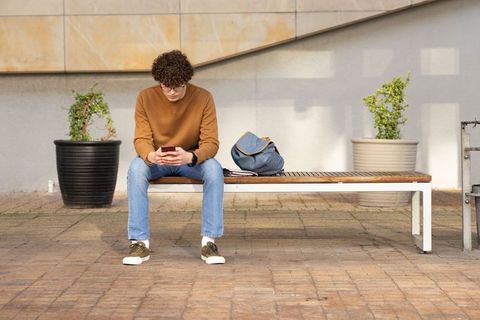 Young student in casual clothes sitting on bench in courtyard holding smartphone with backpack