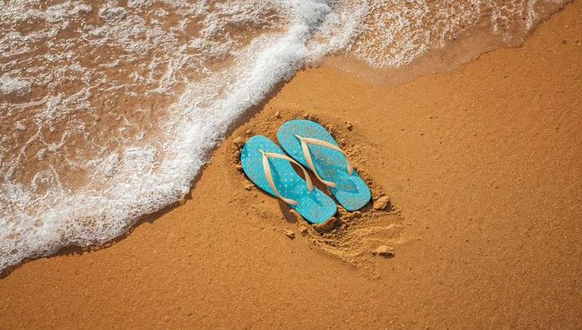 Turquoise flip-flops nestled in golden sand near foamy shoreline, summer beach escape