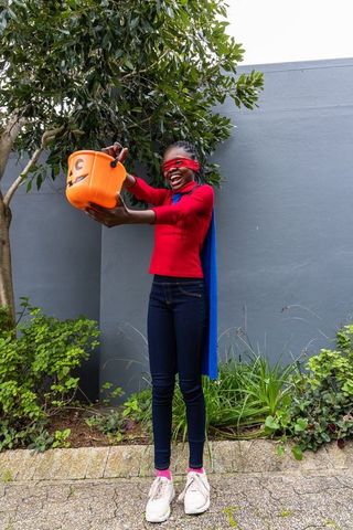 African american teen in superhero cape and blindfold holding halloween pumpkin bucket