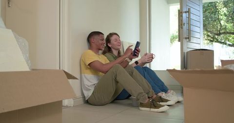 Young couple sitting on floor unpacking boxes, laughing while looking at smartphone