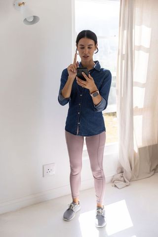 Young woman using smartphone and pointing to smartwatch in bright home wellness lifestyle