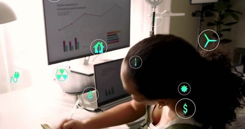 Woman writing at desk analyzing renewable energy charts on dual screens