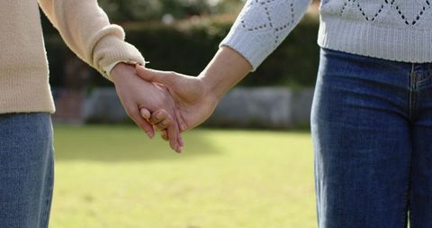Closeup showing two women holding hands on sunlit lawn in cozy knit sweaters and jeans
