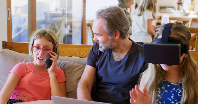Father spending quality time with his daughters. One daughter is using a virtual reality headset, another is talking on the phone, and the father is observing them. Perfect for themes of family bonding, modern technology, and home life.