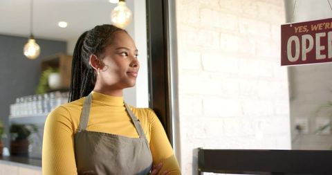 African American Cafe Owner Standing in Mustard Turtleneck and Apron Gazing at OPEN Sign
