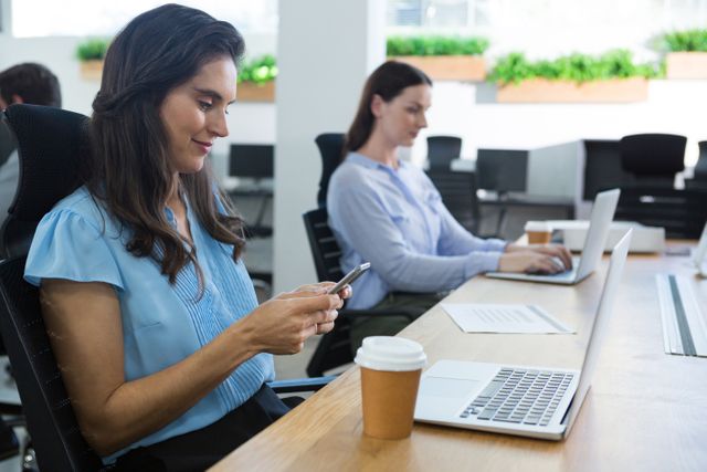 Female executive using mobile phone at office desk with laptop and coffee cup. Colleague working on laptop in background. Ideal for business, technology, and workplace productivity themes.