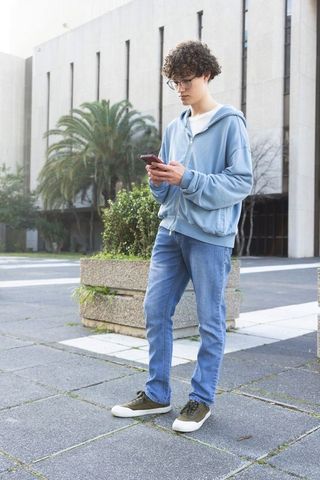 Teen male student standing on campus plaza using smartphone wearing hoodie and glasses