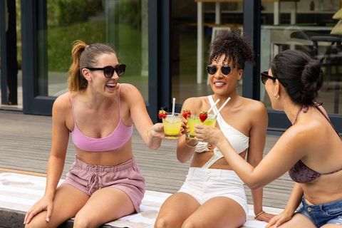 Diverse women clinking cocktails on sunlit deck, laughing and wearing sunglasses