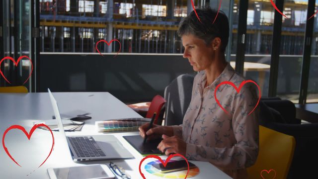 A female graphic designer works attentively with a tablet and laptop in a contemporary office setting, with red heart icons artistically overlaying her workspace. This image conveys themes of creativity, technology, and passion, suitable for illustrating articles or content on digital art, tech innovation with a personal touch, or finding inspiration at work.