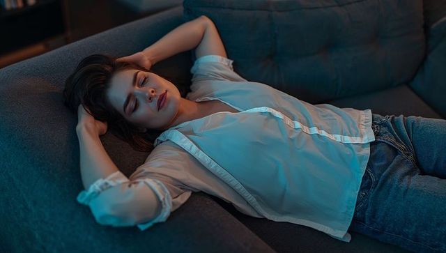 Young woman reclining on sofa in moody ambient light wearing semi-sheer blouse and jeans