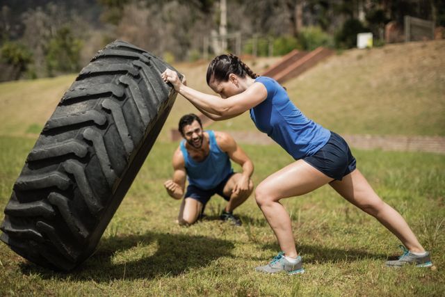 Fit woman flipping a large tire while her trainer cheers her on in an outdoor boot camp setting. Ideal for use in fitness blogs, workout programs, motivational posters, and health and wellness advertisements.