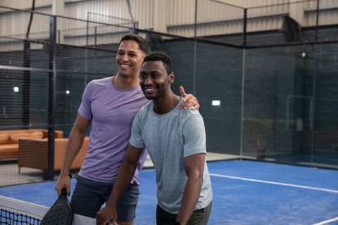 Smiling diverse male friends posing at pickleball net holding paddles on indoor blue court
