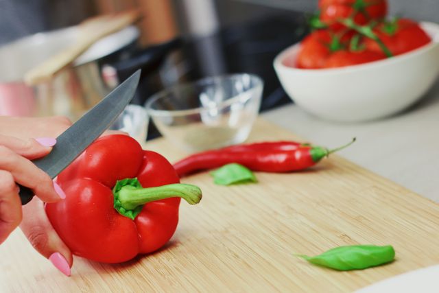 Hands cutting a red bell pepper with a knife on a wooden cutting board in a kitchen. Fresh vegetables including chili and basil leaves are visible. Ideal for articles on healthy eating, recipe blogs, culinary workshops, and kitchen decor.