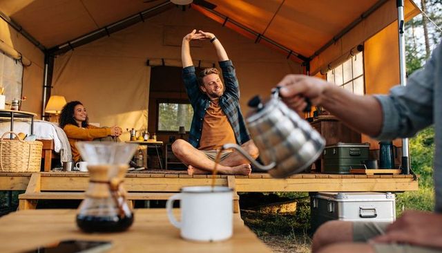 Couple enjoying glamping morning with man stretching on deck and friend pouring coffee