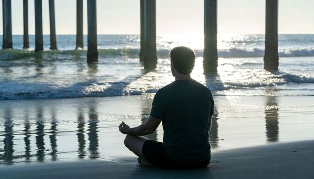 Man meditating on wet sand beneath pier silhouette, ocean reflections at sunset