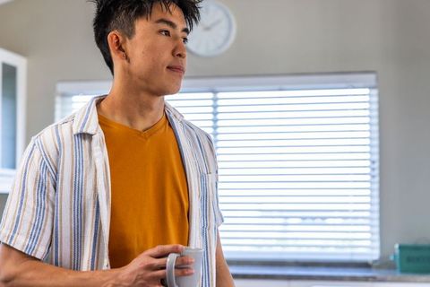 Contemplative asian man standing in bright kitchen holding mug morning coffee